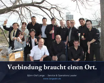 Gruppenfoto in Kyoto: Björn-Olaf Lange kniet im weißen Hemd vorne in der Mitte, um ihn herum Kolleginnen und Kollegen von Kyocera, viele in schwarzen Kyocera-Hemden, mit Bechern in der Hand unter einem Baum. Im Banderolentext: Verbindung braucht einen Ort.